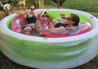 Two women joyfully celebrating birthday by sitting on pink floats in kiddie pool and drinking margaritas. 